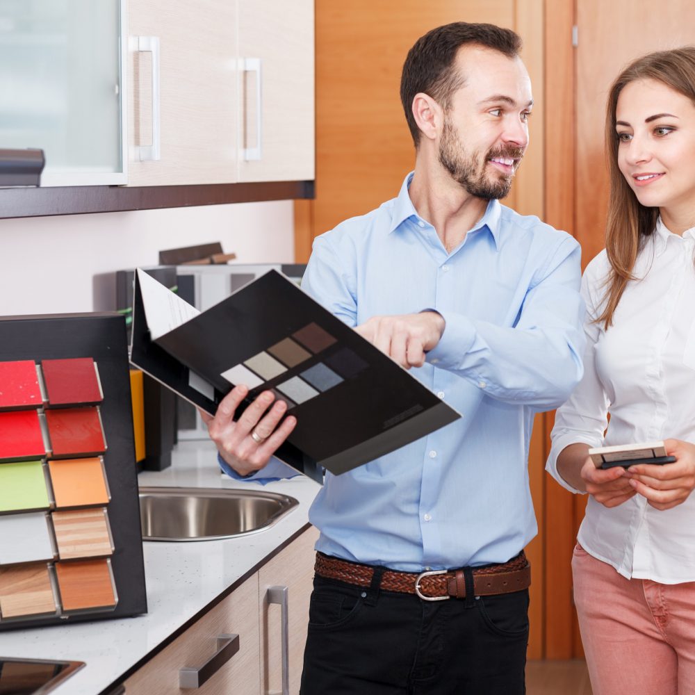 Smiling polite salesman helping young woman to choose materials for kitchen furniture in shop